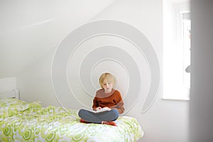 Smart preteen boy sitting on bed and enthusiastically reading interesting book. Kid reader enjoying interesting stories, reading