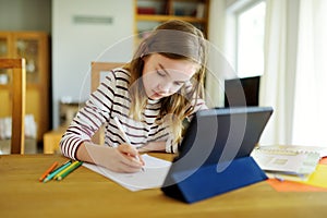 Smart preteen schoolgirl doing her homework with digital tablet at home. Education and distance learning for kids. Homeschooling