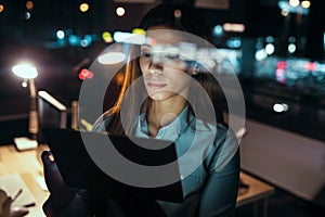 Smart pretty business woman taking a break while cheaking her digital tablet standing in the office at night