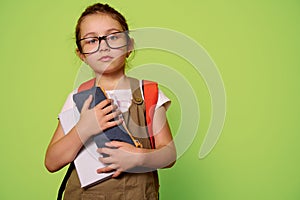 Smart primary school student girl in glasses, holding book and pencil case, looking at camera isolated green background