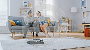 Smart Robot Vacuum Cleaner Sucking Up Dust from a Carpet. Beautiful Couple is Sitting on a Sofa an