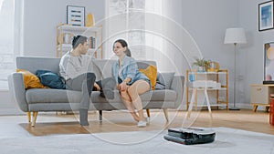 Smart Robot Vacuum Cleaner Sucking Up Dust from a Carpet. Beautiful Couple is Sitting on a Sofa an