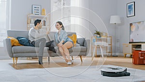 Smart Robot Vacuum Cleaner Sucking Up Dust from a Carpet. Beautiful Couple is Sitting on a Sofa an