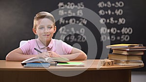 Smart schoolboy smiling to camera after solving task, exercises written behind