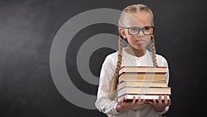 Smart schoolgirl in eyeglasses hardly holding heap of books against blackboard