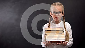 Smart schoolgirl in eyeglasses hardly holding heap of books against blackboard