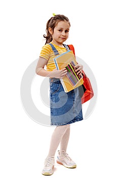 Smart schoolgirl in yellow t-shirt and denim sundress, carrying backpack and school supplies, smiles on white background