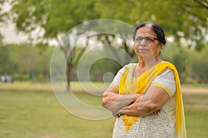 Smart senior north Indian woman standing, posing for the camera with hands crossed in a park wearing yellow salwar kameez in