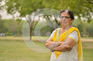 Smart senior north Indian woman standing, posing for the camera with hands crossed in a park wearing yellow salwar kameez in photo