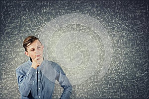 Smart teenage boy thinking, keeps hand under chin, stands in front of a blackboard solving hard mathematics calculation and