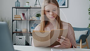 Smart teenage girl reading book turning pages sitting at table at home concentrated on studies