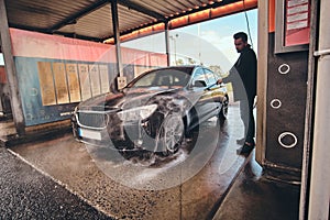 Smart trendy man in jeans and blaser is washing his own car at car washing station