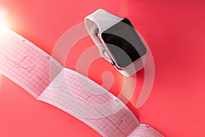 Smart watch on white sports silicone band, ECG diagram paper on pink coral background. Cardiogram medical examination