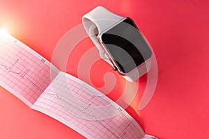 Smart watch on white sports silicone band, ECG diagram paper on pink coral background. Cardiogram medical examination