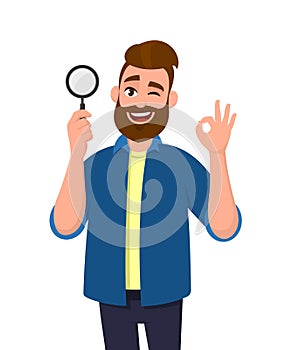 Smart young man holding magnifying glass and gesturing okay/OK sign while winking eye. Deal, good, agree, search, find, discovery.