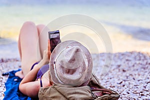 Smartphone on the beach: beautiful young girl with straw hat in bikini is lying on the pebble beach, vacation in italy