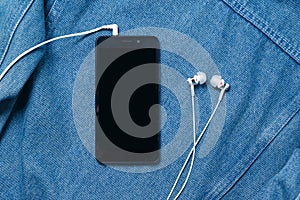 Smartphone with black case and a blank white screen and plugged in headphones ear buds on the background of denim