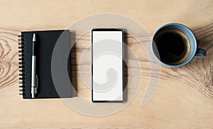Smartphone with blank white mock up screen, notebook and coffee cup on wooden office desk.