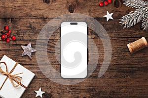 Smartphone with blank white screen on rustic wooden background, surrounded by Christmas decorations, pine branches, and festive