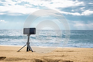 A smartphone camera on a tripod in the foreground stands on the seashore