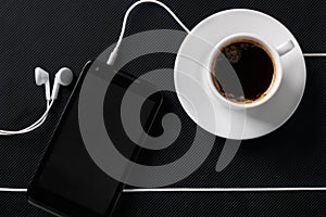 Smartphone with earphones and cup of coffee on black textured surface. Flat lay
