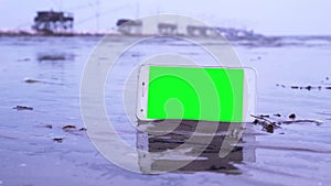 Smartphone with green screen on the beach, bathed by sea water