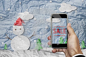 Smartphone on hand with `SALE` on screen, with crumpled paper cut snowman in winter, Christmas shopping sale