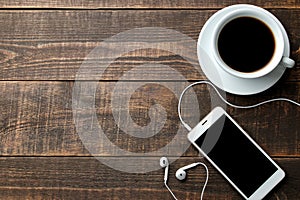 Smartphone with headphones and a cup with coffee on a brown wooden table. view from above