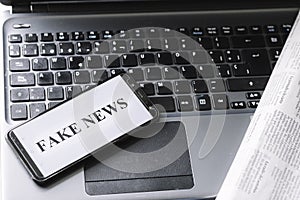 Smartphone with the phrase fake news written on the screen on a laptop, newspaper and magnifying glass