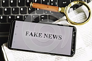 Smartphone with the phrase fake news written on the screen on a laptop, newspaper and magnifying glass