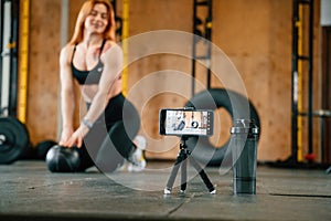 Smartphone is on the tripod, live stream, capturing video. Beautiful strong woman is in the gym