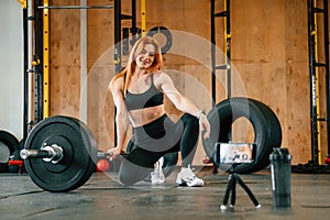 Smartphone is on the tripod, live stream, capturing video. Beautiful strong woman is in the gym