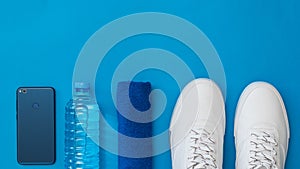 Smartphone, water bottle, towel and white sports shoes on blue background. Sports style. Flat lay.