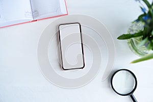 Smartphone with white blank screen. Notebook, magnifier and flowers on table