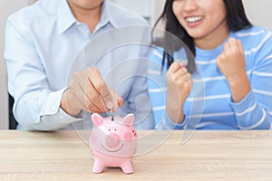 Smile couple putting a coin into a pink piggy bank on wooden desk - save money for the future.