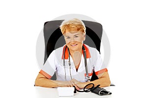 Smile elderly female doctor or nurse sitting behind the desk with bloog preasure gauge