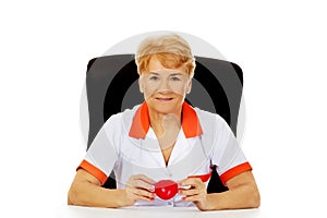 Smile elderly female doctor or nurse sitting behind the desk and holds heart toy