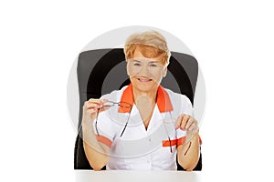 Smile elderly female doctor or nurse sitting behind the desk and holds two pair of glasses