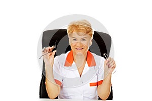 Smile elderly female doctor or nurse sitting behind the desk and holds two pair of glasses