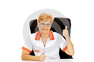 Smile elderly female doctor or nurse sitting behind the desk and shows thumb up