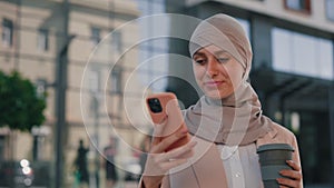 Smiled arabian business woman wearing hijab standing near the business centre using smartphone, texting with friends