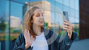 Smiled caucasian woman using smartphone, video calling her friends, waving at the camera, wearing ear phones, telling