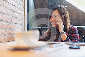 A smiley beautiful Asian woman sitting on sofa , using and talking on smart phone in modern cafe