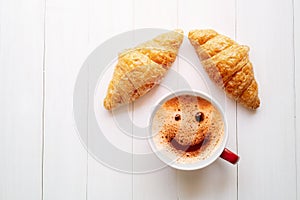 Smiley face foam coffee with croissant on a white wooden table