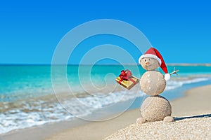 Smiley sandy snowman at beach in christmas hat with golden gift.