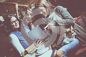 Smiley three girls students in cafe using digital tablet and having pet cat.