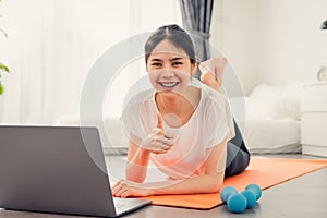 Smiley young Asian woman on a yoga mat and raises her hand to show like pose on the bedroom. Watch exercise online via the laptop.