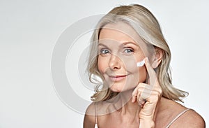 Smiling 50s middle aged mature woman applying cream on face  on white.