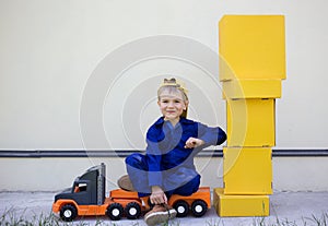 smiling 6 year old boy in blue uniform is sitting on big toy truck next to a lot of yellow cardboard boxes