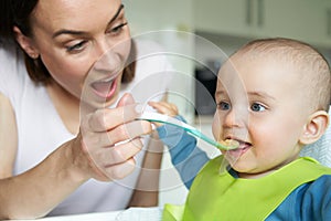 Smiling 8 month Old Baby Boy At Home In High Chair Being Fed Solid Food By Mother With Spoon